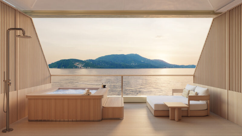 Modern yacht deck with a hot tub, outdoor shower, and lounge seating, overlooking calm water and a distant green island at sunset.