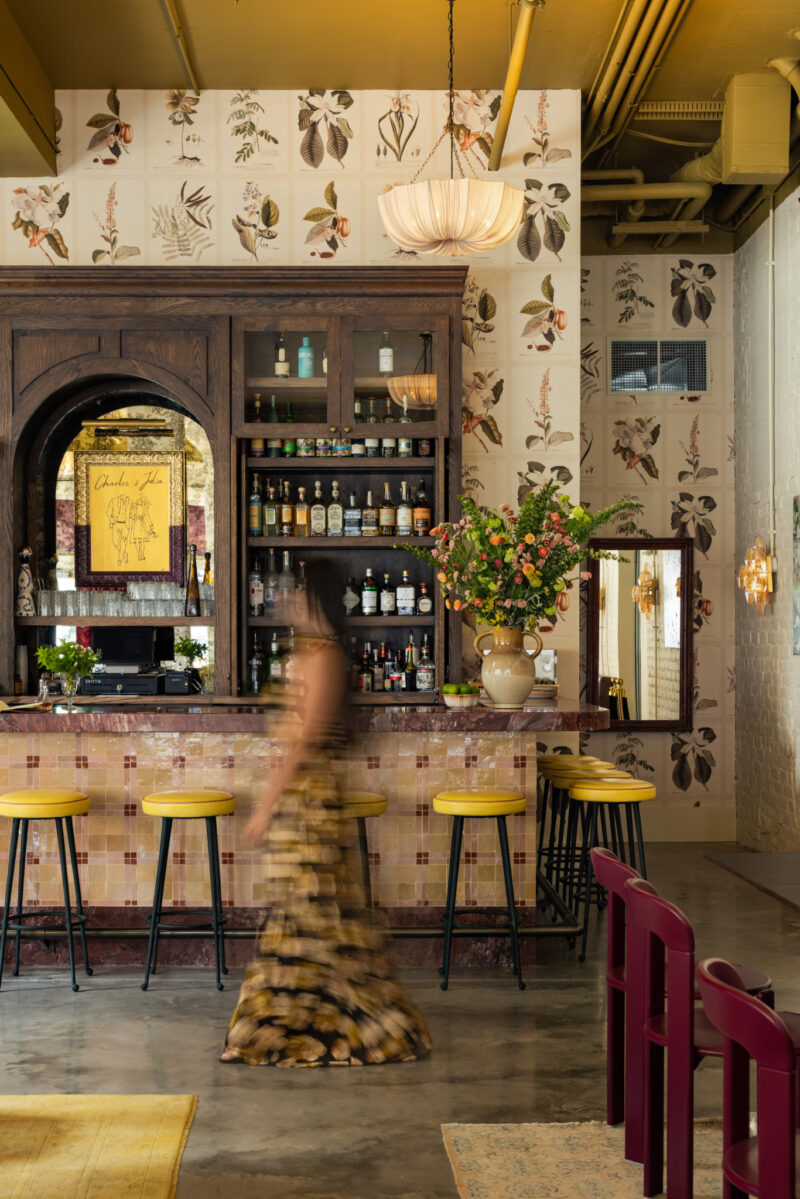 Una mujer con un vestido de flores pasa por el bar Charles & Julia, decorado con taburetes amarillos, grandes arreglos florales y botelleros, y decorado con papel tapiz botánico.