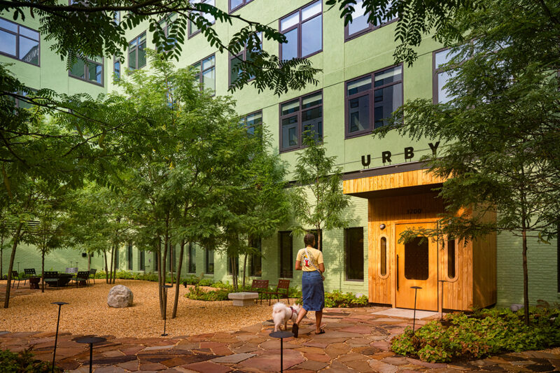Un hombre y un perro se encuentran en un patio ajardinado en las afueras de Fishtown Urby, un edificio de apartamentos verde con una entrada de madera etiquetada "Vaya, ¡vaya!