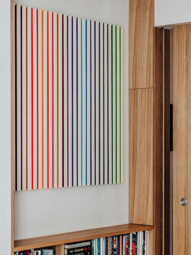 En una habitación contemporánea, un cuadro de franjas verticales espaciadas uniformemente de diferentes colores cuelga de una pared blanca encima de una estantería.