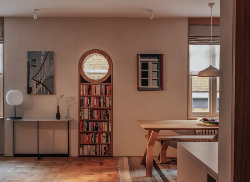 Moderno salón-comedor con ventana redonda sobre una estantería, mesa de comedor de madera, arte en la pared y mesa consola con lámpara y jarrón.