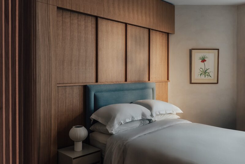 Una cama cuidada con ropa de cama blanca junto a una mesita con una lámpara, una cabecera con paneles de madera y arte floral enmarcado colgado en la pared.