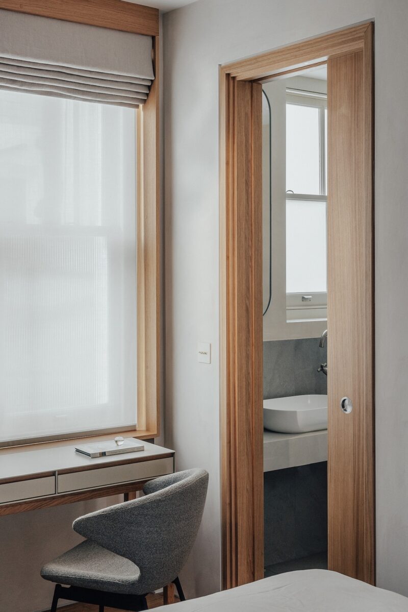Dormitorio moderno y minimalista con escritorio, silla gris y puerta corredera de madera que conduce al baño con lavabo y ventana.