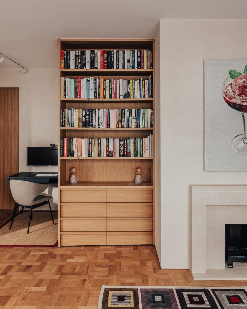 Entre la pequeña zona de despacho y la chimenea del moderno salón se encuentra una estantería de madera repleta de libros y dos objetos decorativos.