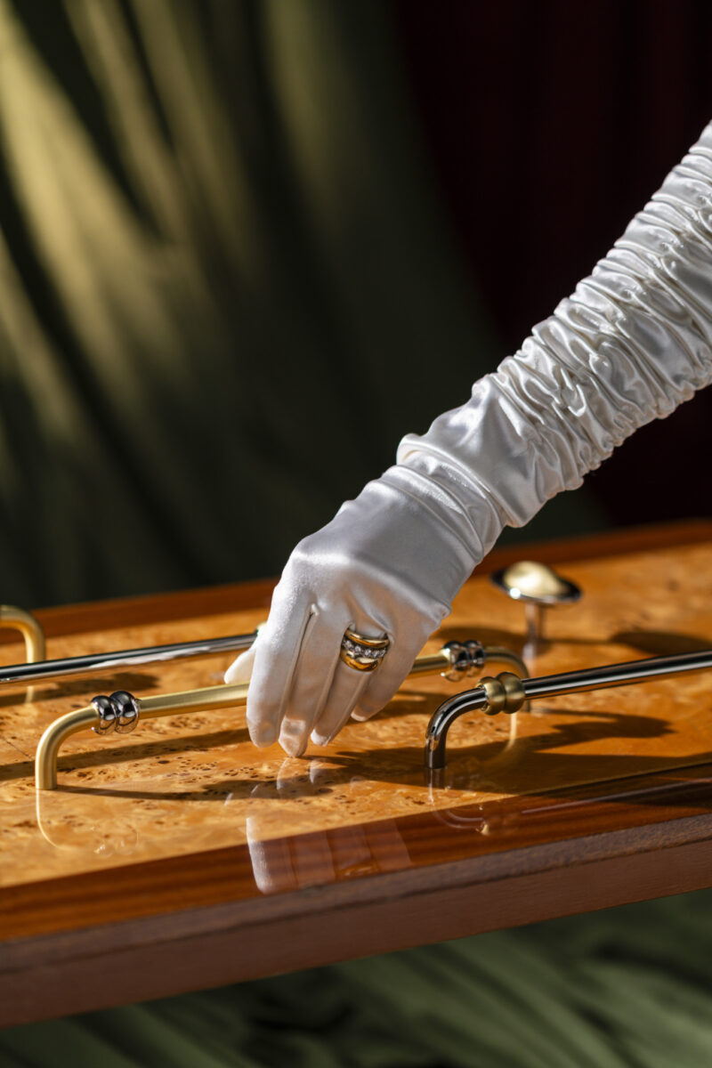 A person wearing a white ruched glove and a gold ring is touching a polished wooden surface with brass handles and rods.