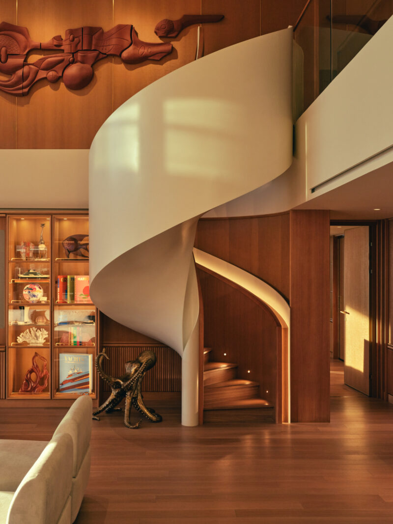 Curved white spiral staircase in a warm-toned interior, with bookshelves, art, and sculptural decor surrounding the modern wood and glass space.