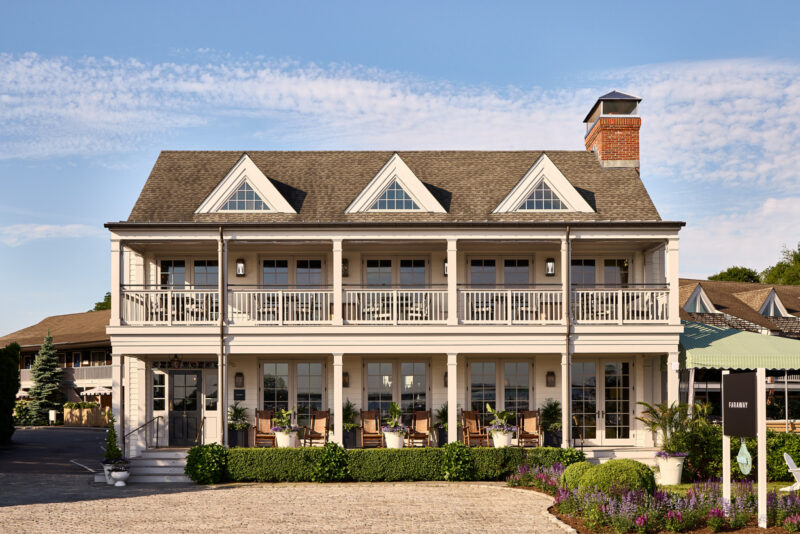 Two-story building with a large front balcony, multiple dormer windows, and a central chimney, surrounded by landscaping under a clear sky.