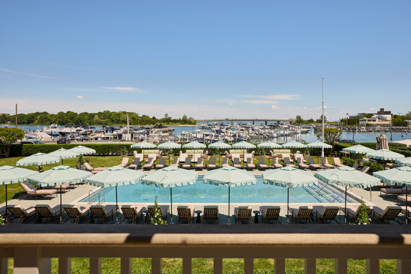 A swimming pool surrounded by lounge chairs and green umbrellas, overlooking a marina with boats docked under a clear blue sky.