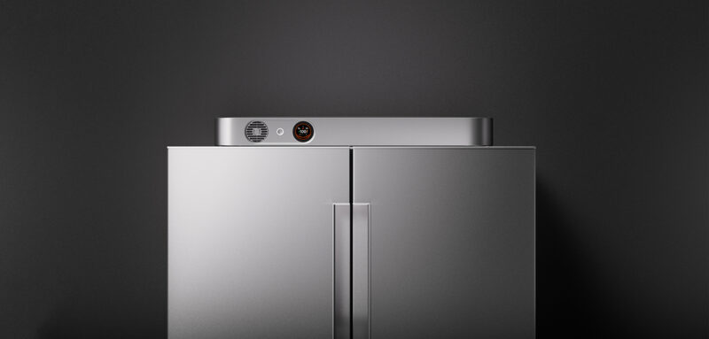 A metallic double-door refrigerator stands against a dark gray wall, featuring a small control panel and vent on the top edge.