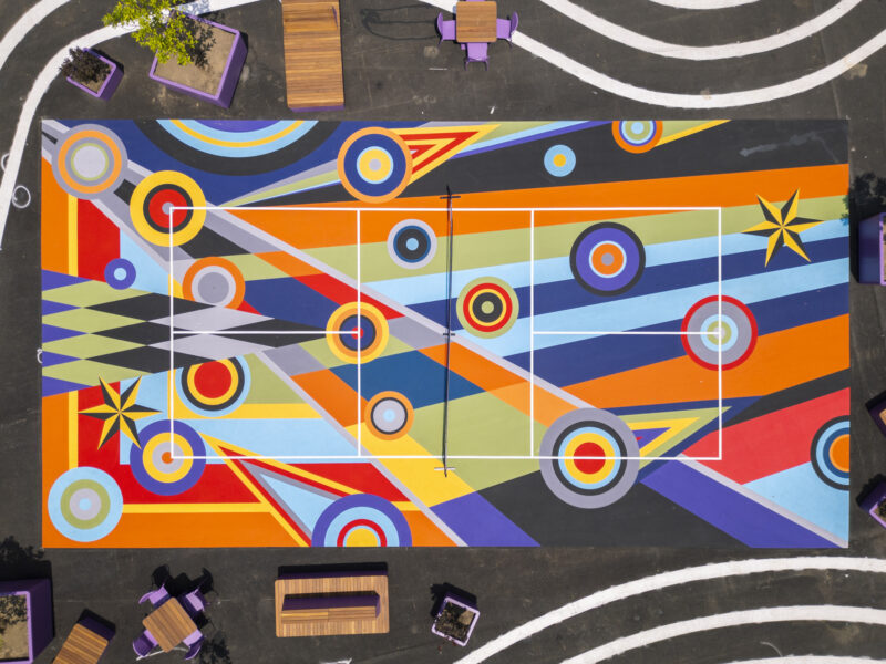 Una vista aérea del campo deportivo de colores brillantes con patrones geométricos inspirados en la Geometría del Juego de Sharmistha Ray, rodeado de bancos y plantas.