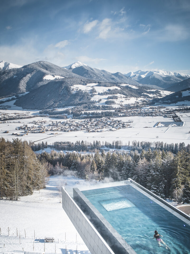 La piscina al aire libre, parte de una ampliación del spa del Hotel Hubertus, se extiende sobre un paisaje nevado con montañas y pueblos al fondo. Bajo el cielo despejado, una persona nada en la piscina.