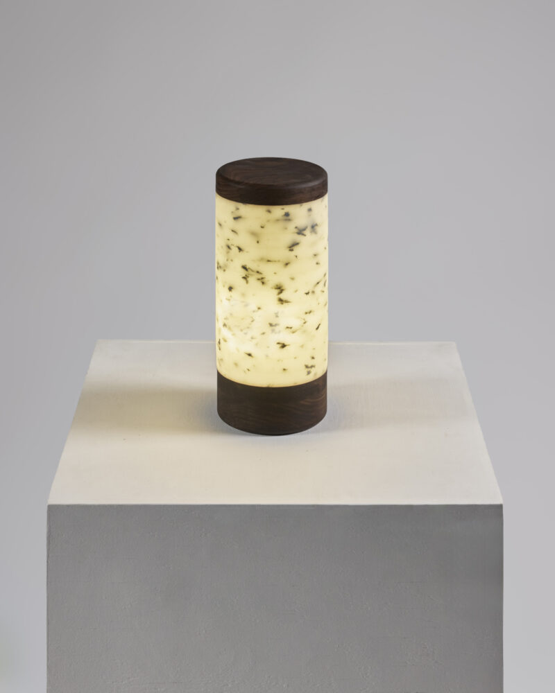 A cylindrical lamp with a dark top and base, and a translucent white body with speckled details, sits on a plain white pedestal against a neutral background.