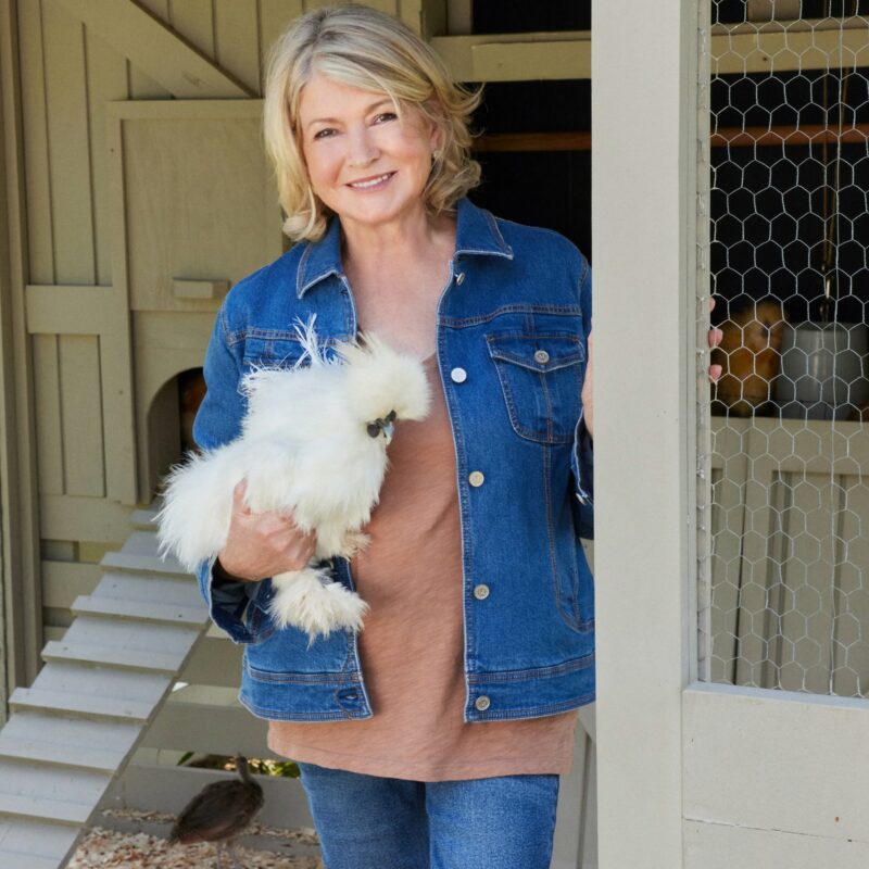 De pie junto a un gallinero, una mujer con una chaqueta vaquera sostiene un pollo blanco esponjoso en sus manos y sonríe a la cámara, encarnando la combinación característica de calidez y estilo de Martha Stewart.