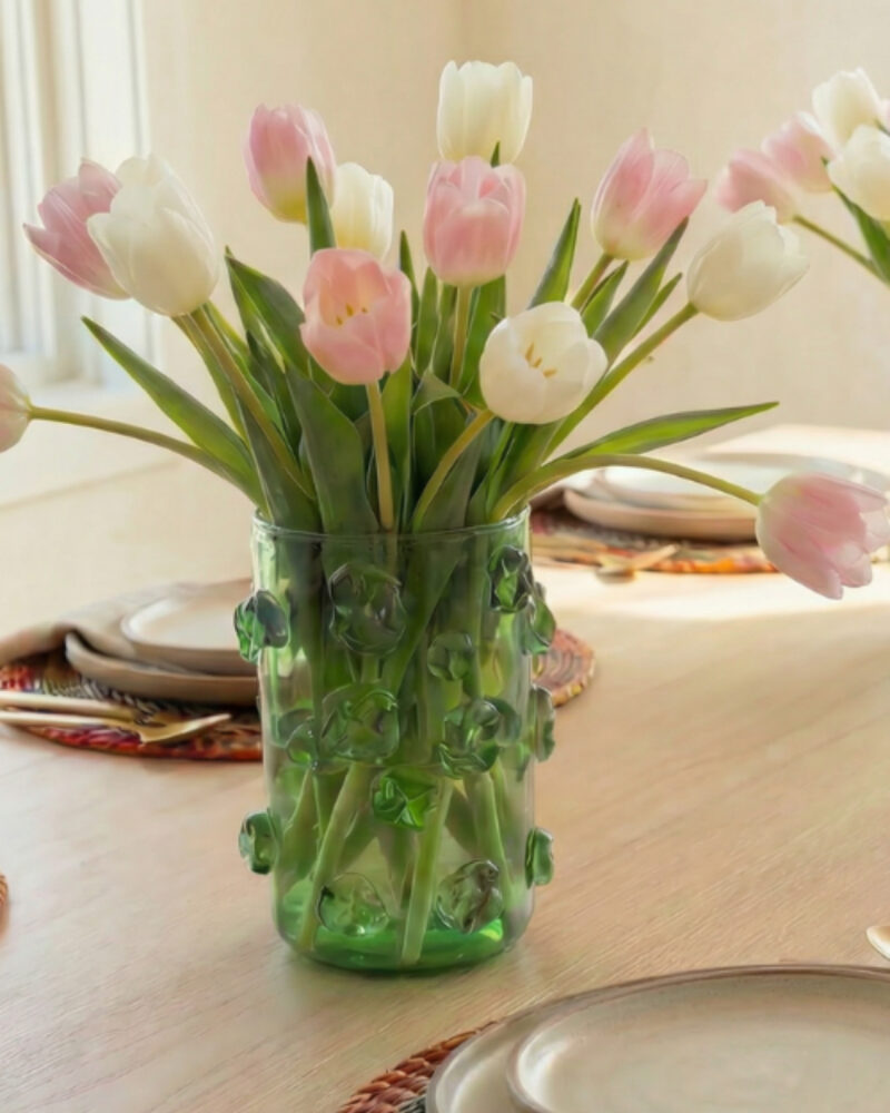 A green glass vase with white and pink tulips is placed on a wooden dining table set with plates and cutlery, bringing a touch of modern African decor to the space.
