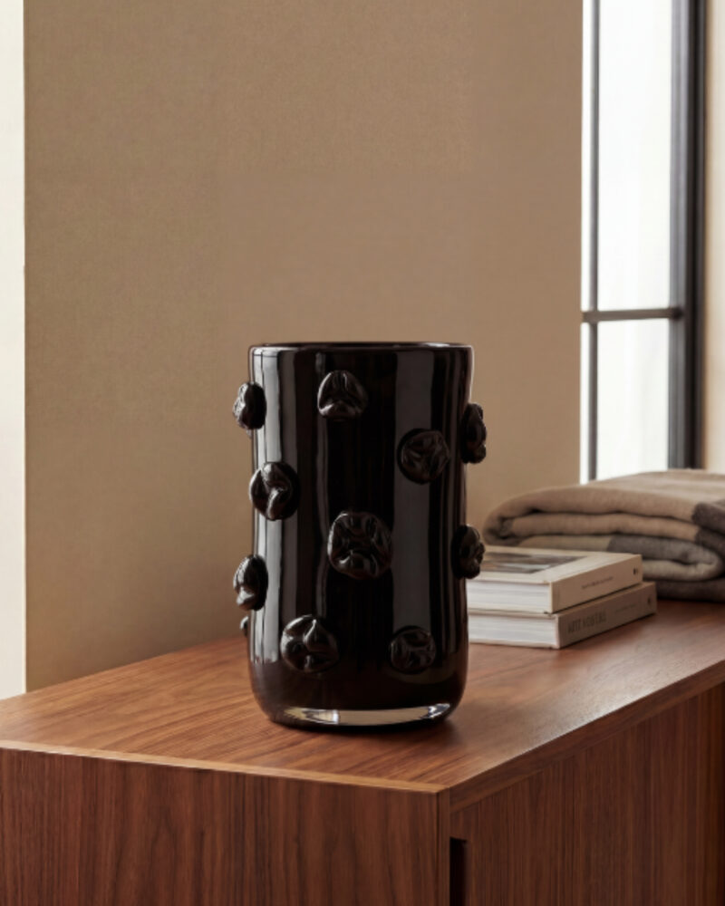 A glossy black vase with raised circular knobs, inspired by modern African decor, sits on a wooden sideboard next to a small stack of books and folded blankets near a window.