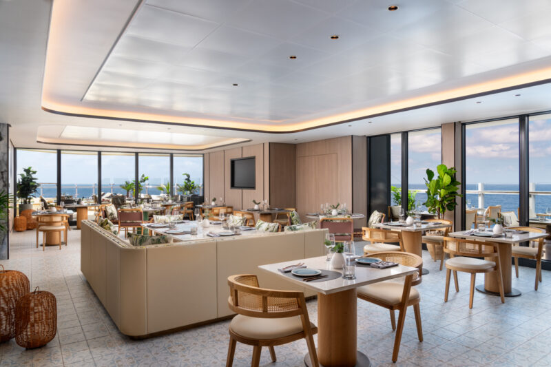 Modern restaurant dining area with wooden tables and chairs, set for service, large windows offering ocean views, and natural light filling the space.