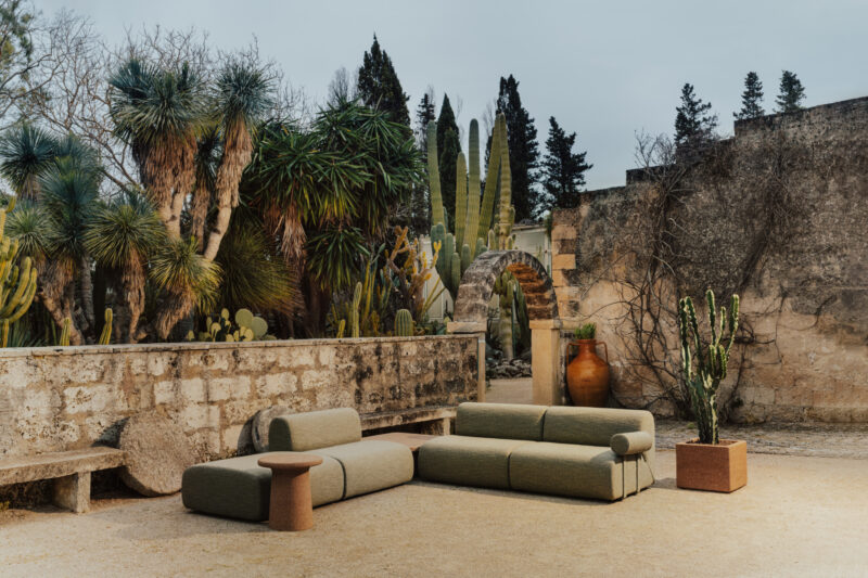 La zona de estar al aire libre cuenta con un sofá modular, una mesa auxiliar y cactus en macetas, colocados contra una pared de piedra y rodeados por una variedad de plantas y árboles del desierto.