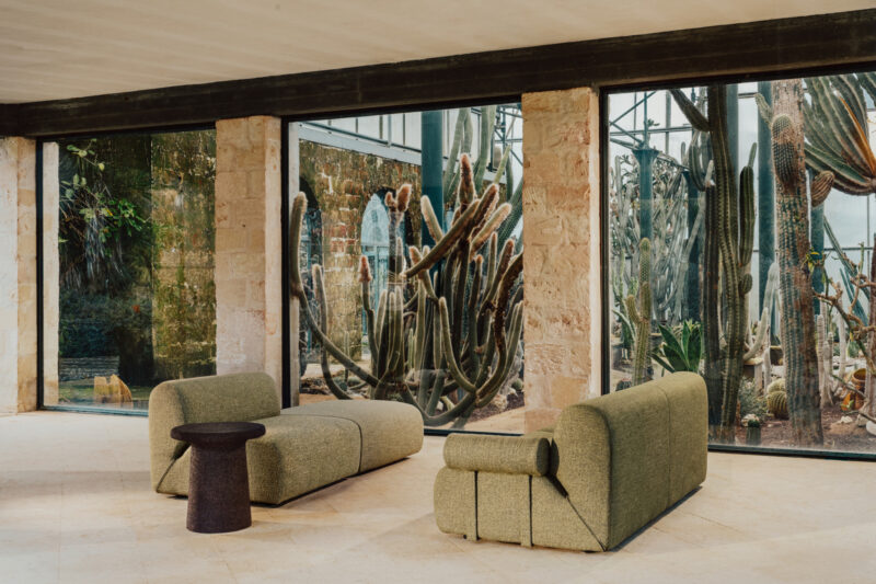 Salón moderno con dos sofás verdes y una pequeña mesa redonda en una habitación con grandes ventanales con vistas a un jardín de cactus.
