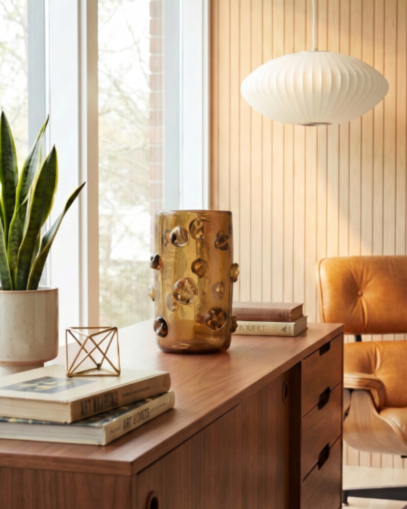 A modern office with wooden desk, brown leather chair, potted plants, books, amber vase and a white pendant lamp - enhanced by subtle touches of modern African decor - in vertical wood paneling and large windows.