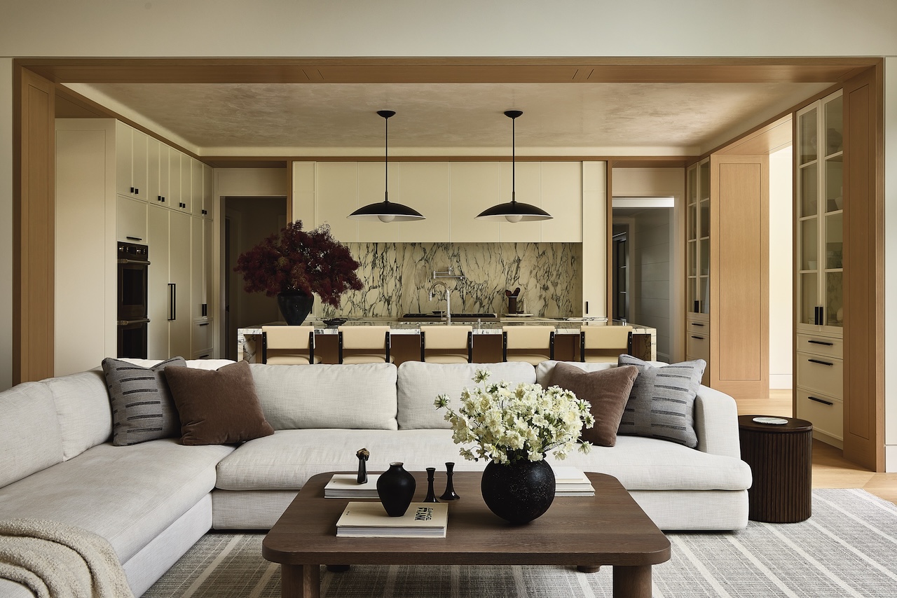 Modern open-concept living room and kitchen with neutral tones, marble backsplash, pendant lights, a sectional sofa, and a wooden coffee table with flowers and decorative items.