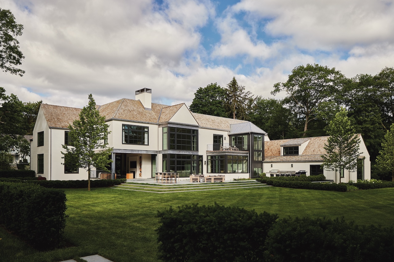 Modern two-story house with large windows, white exterior, and attached garage. The lawn is well-manicured, and there are mature trees and outdoor seating in the backyard.