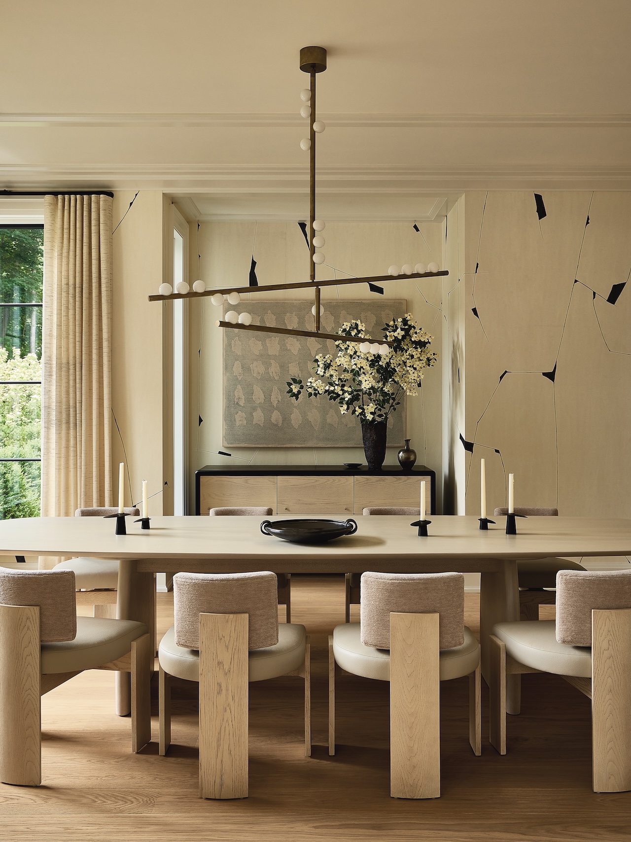Modern dining room with light wood table and chairs, minimalist decor, geometric lighting fixture, artwork, vase with white flowers, and large window with beige curtains.