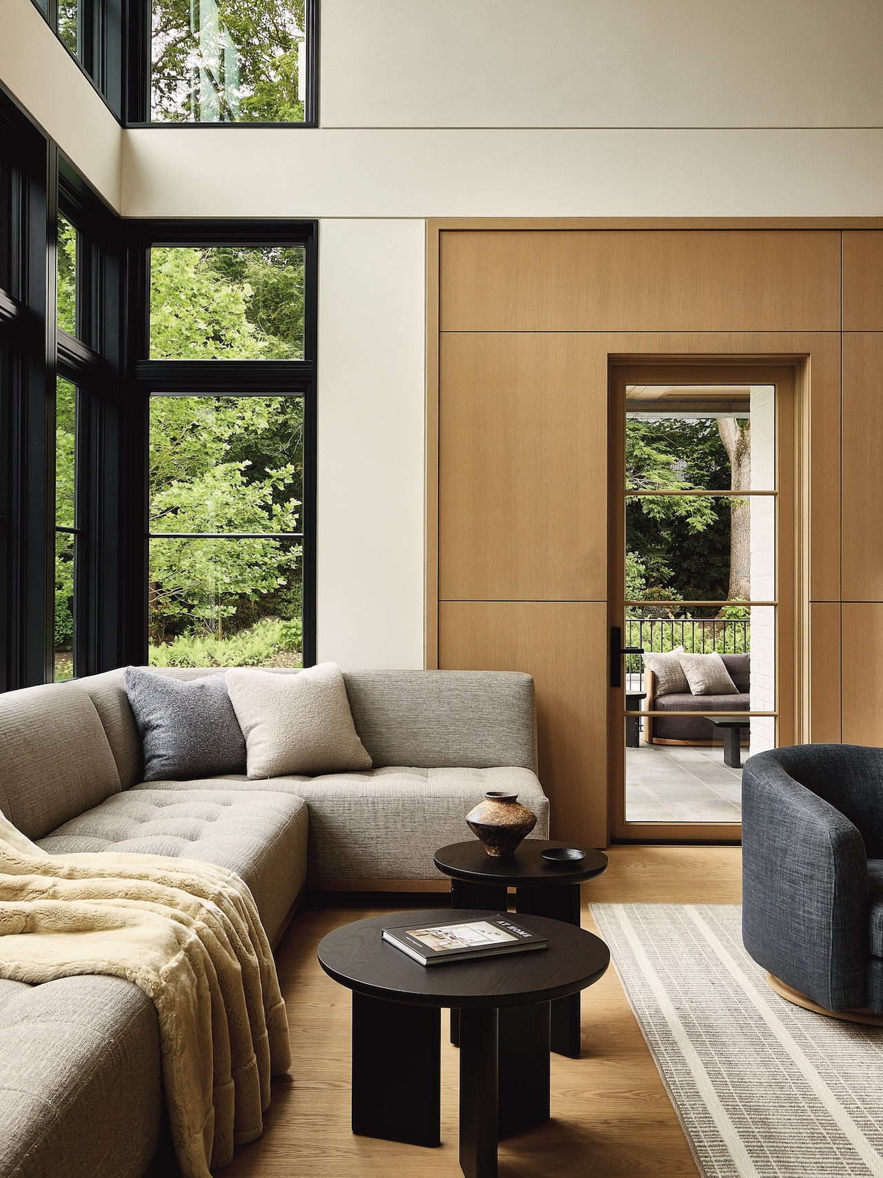 Modern living room with large windows, light wood walls, a gray sectional sofa, a dark armchair, and a round coffee table. A door opens to a patio with trees visible outside.