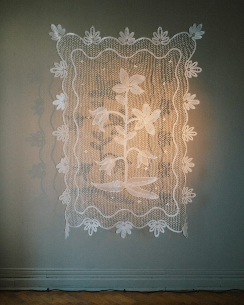 A white lace textile with a floral pattern hangs on a gray wall, bordered with decorative leaves and a scalloped edge—a delicate touch reminiscent of Baum und Pferdgarten’s signature elegance.