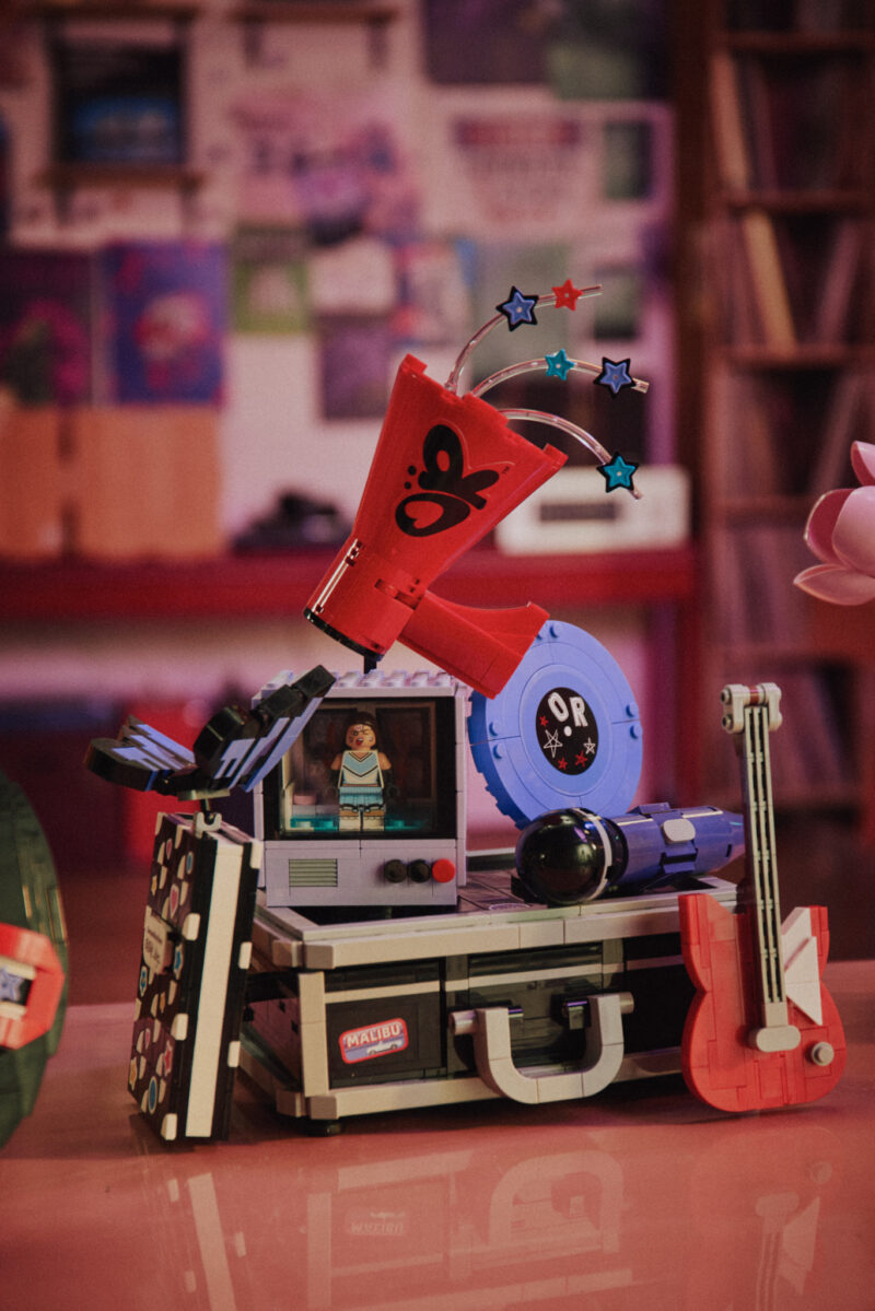 A LEGO model featuring a suitcase, a red guitar, a blue record player, a TV screen with a figure, and a large red megaphone with stars on springs.