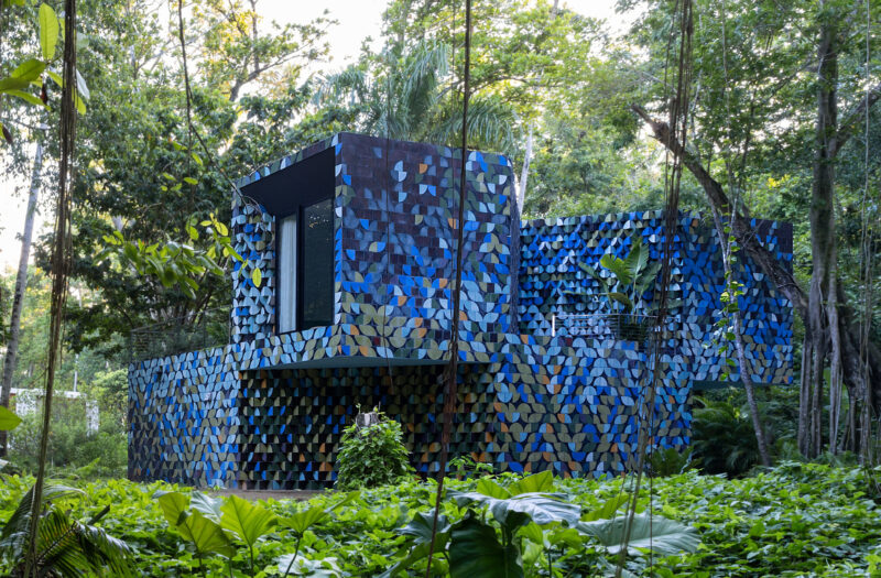 Modern building with a facade of blue and purple tiles stands amid dense green foliage and trees in a forested area.