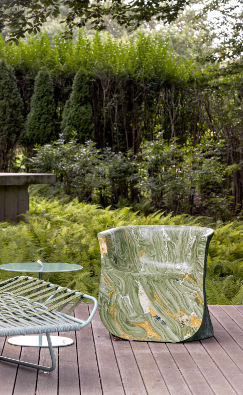 A green and white marbled chair and a metal table sit on a wooden deck surrounded by lush ferns and dense greenery.