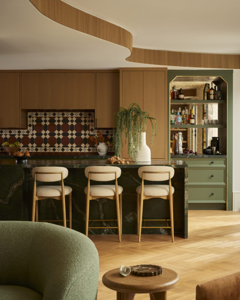 Modern kitchen with wood cabinets, patterned tile backsplash, three bar stools, green accents, a curved ceiling detail, and a bar area with bottles and glassware in the background.