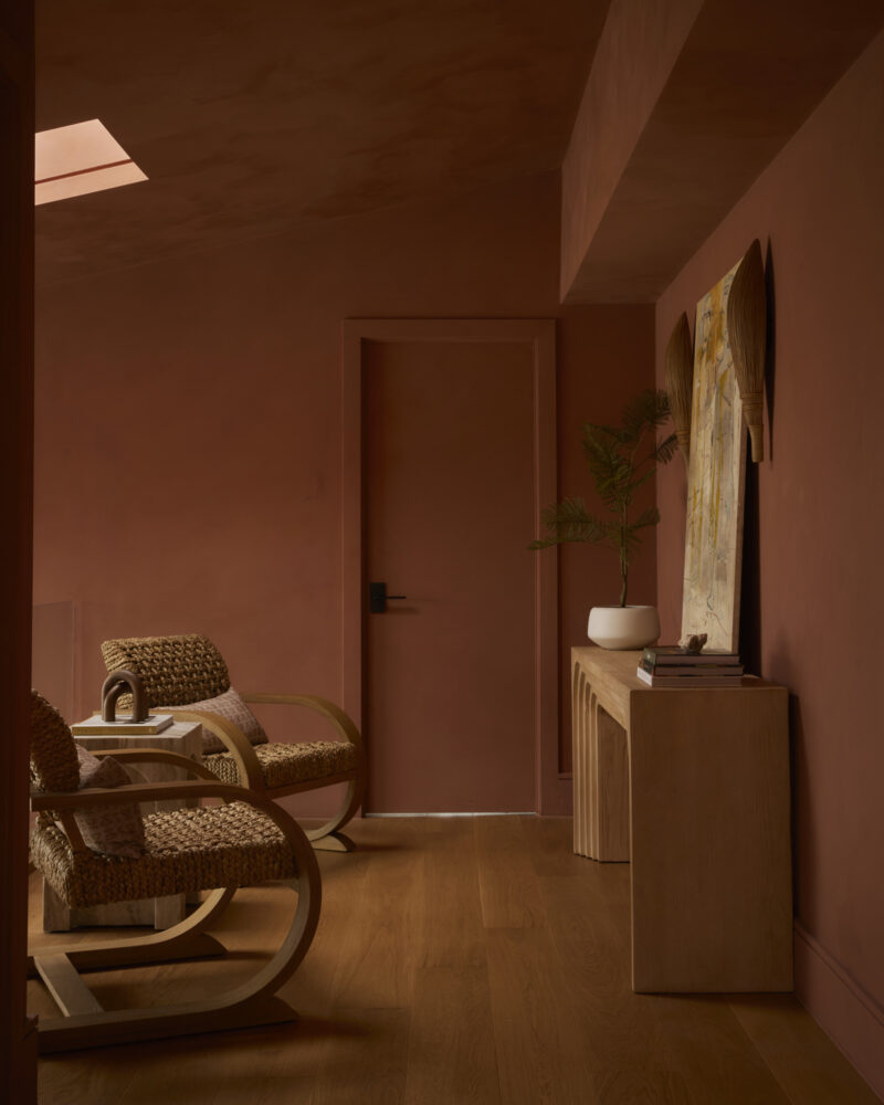 A room with two woven chairs, a small side table, a console with a potted plant, and artwork on a wood floor, all in warm brown and terracotta tones.