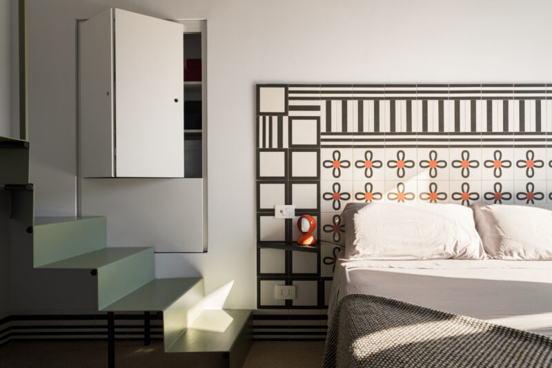 Minimalist bedroom with patterned headboard, white bed, floating glass stairs, open wall cabinet, and sunlight casting shadows on the steps and wall—an elegant retreat inspired by the serene ambiance of La Casa Olive e Capperi.