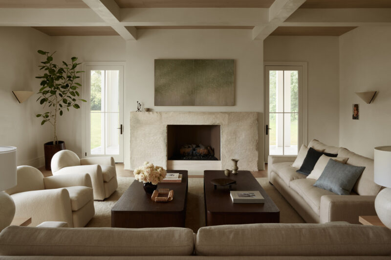 Neutral-toned living room with two sofas, two armchairs, a stone fireplace, wooden coffee tables, a potted plant, and minimal decor. Light streams in from two glass doors.