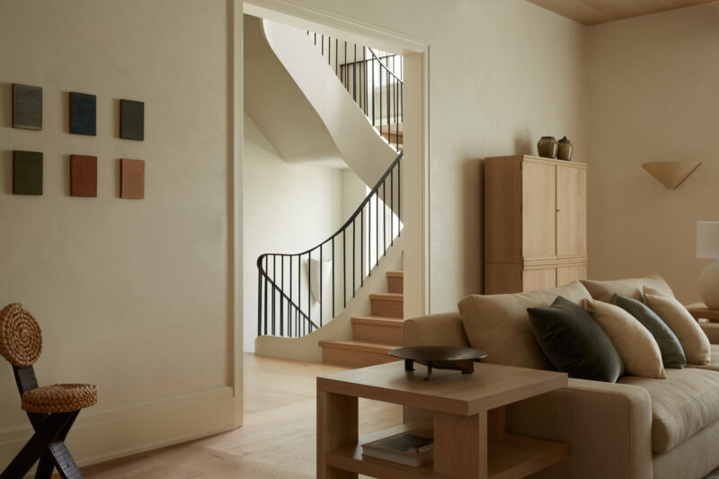 Modern living room with beige sofa, black and wooden accents, wall art, and a view of a staircase with a black railing in the background.