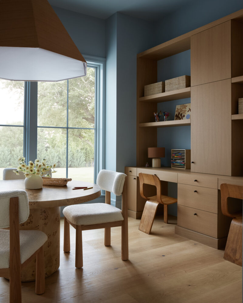 A modern home office with wood furniture, built-in shelves, two desks, two chairs, a round table, and large window letting in natural light.