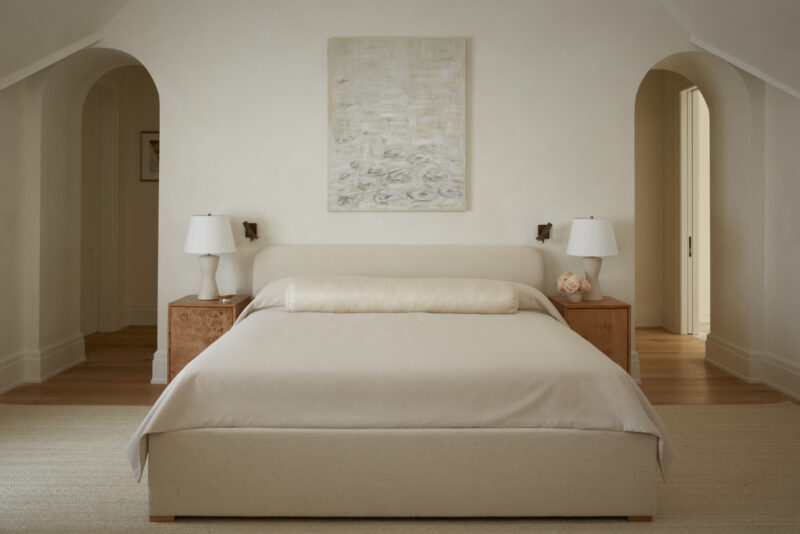 A minimalist bedroom with a beige bed, two wooden nightstands with white lamps, abstract art above the bed, and two arched doorways on either side.