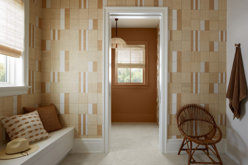 A hallway with patterned beige walls leads to a room with brown walls, wicker chair, window, and hanging light. Cushioned bench and towel hang beside the doorway.