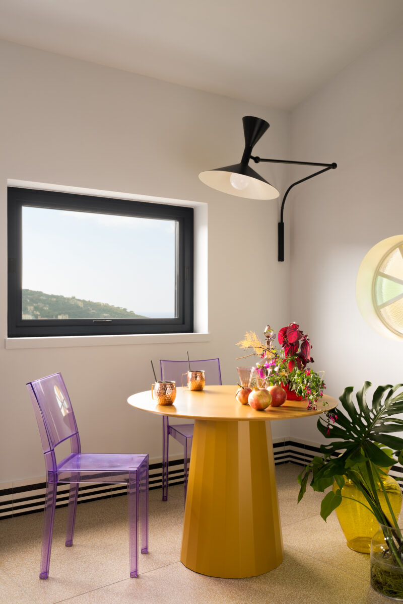 A round yellow table with drinks and fruit is surrounded by two transparent purple chairs in a bright room with modern decor and a wall-mounted lamp, evoking the vibrant and inviting atmosphere of La Casa Olive e Capperi.