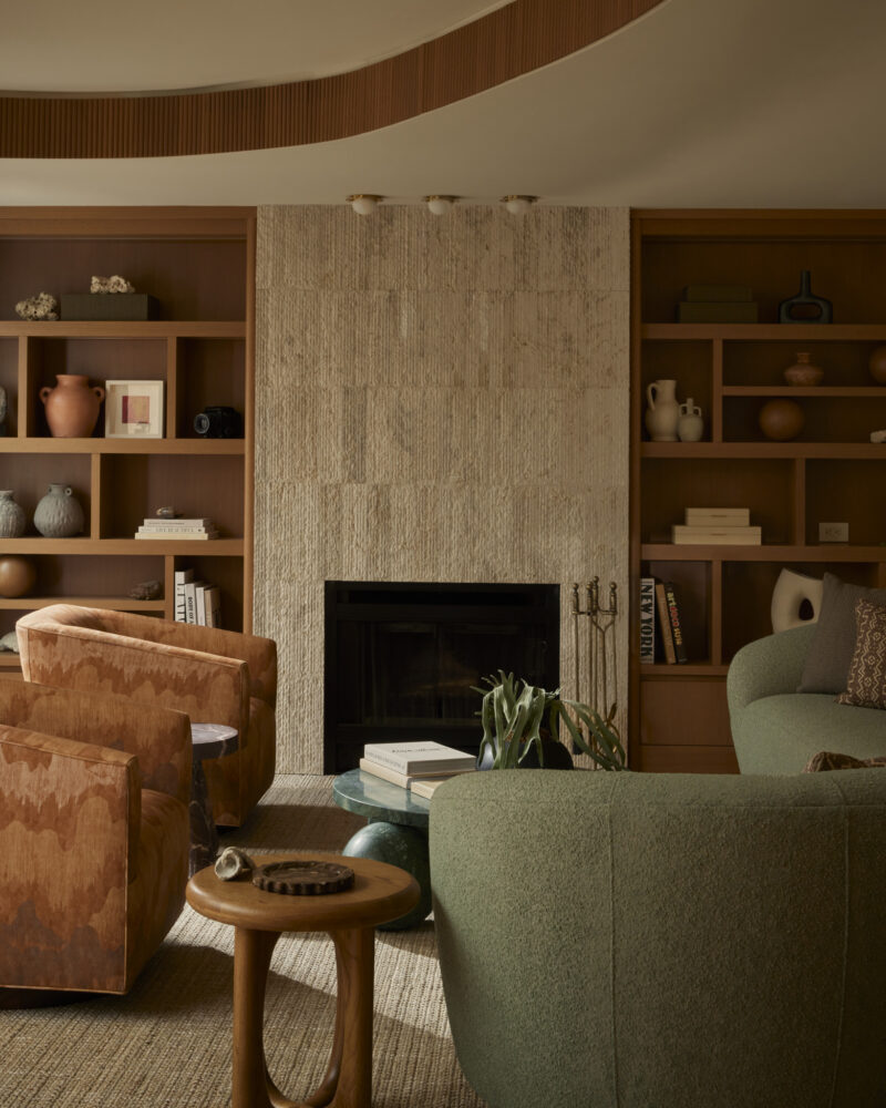 A living room with a stone fireplace, built-in shelves, tan armchairs, a green sofa, and a round side table. Neutral tones and decorative vases create a warm, modern atmosphere.