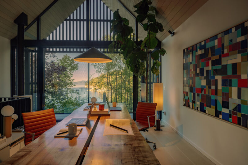 Modern home office with two desks, orange chairs, large windows showing trees and a lake at sunset, a tall plant, and a colorful abstract painting on the wall—this inviting space evokes the spirit of a Little Giant forest studio.