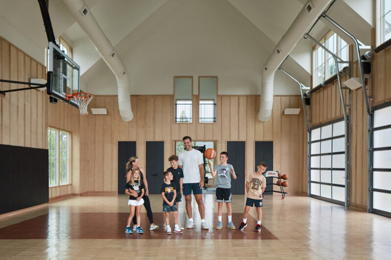 Six children and one adult stand together on a basketball court inside a large indoor gym with wooden walls and high ceilings.