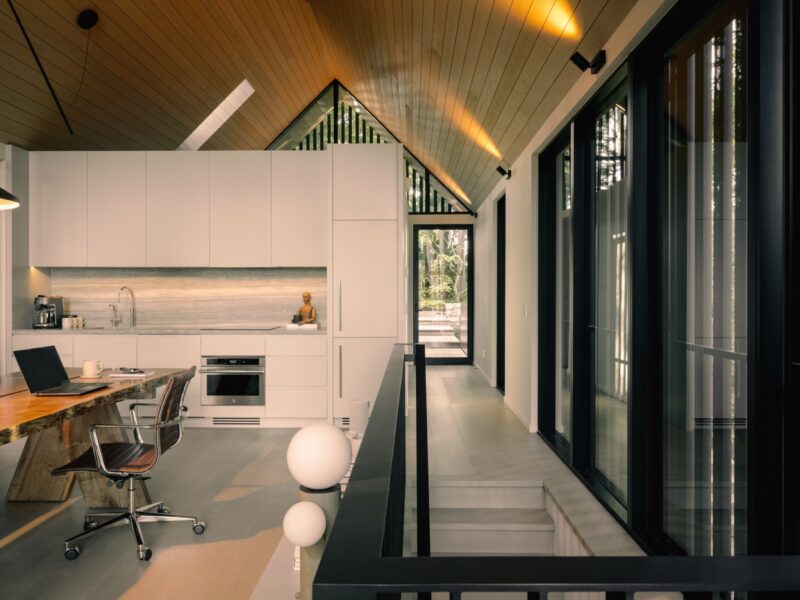 Modern kitchen with white cabinets, built-in appliances, a wooden dining table, and an office chair beside large windows and a sloped wooden ceiling—reminiscent of the cozy creativity found in a Little Giant forest studio.