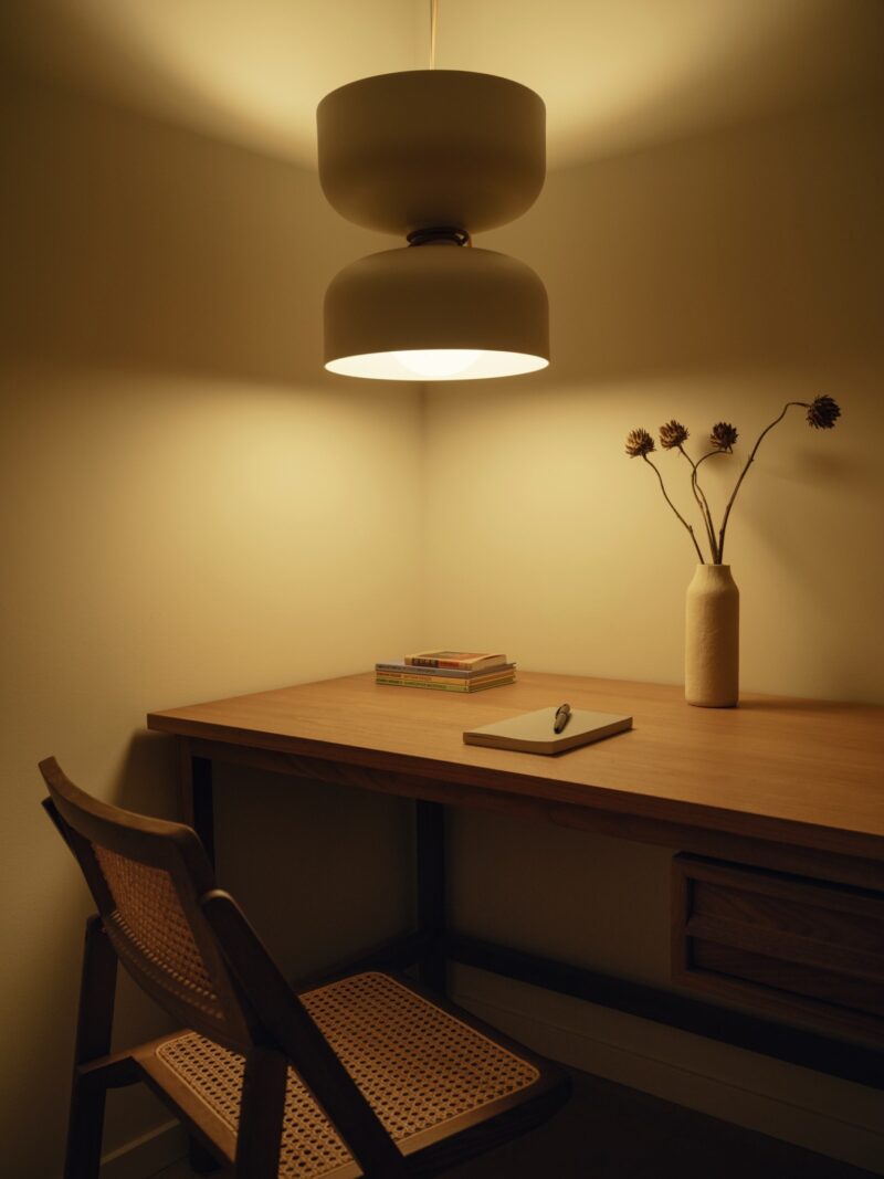 A wooden desk with a notebook, pen, books, and a vase with dried flowers rests under a modern pendant lamp, creating the warm ambiance of a Little Giant forest studio. A woven chair is positioned at the desk.