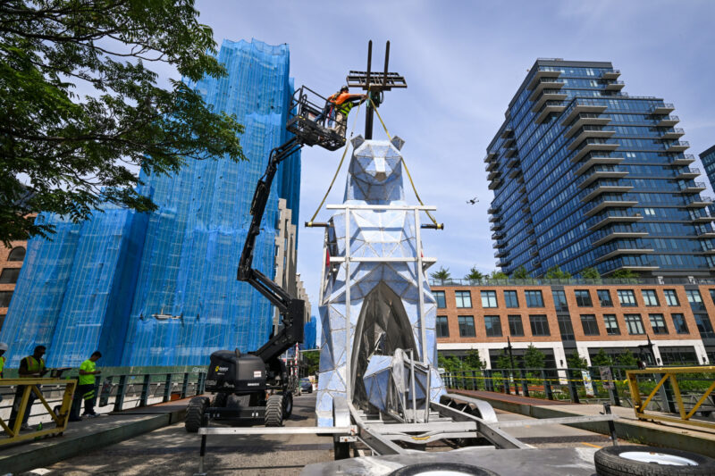 Workers on a lift install Jen Lewin’s The Ursas, a striking geometric animal sculpture, in an urban setting surrounded by high-rise buildings and construction scaffolding.