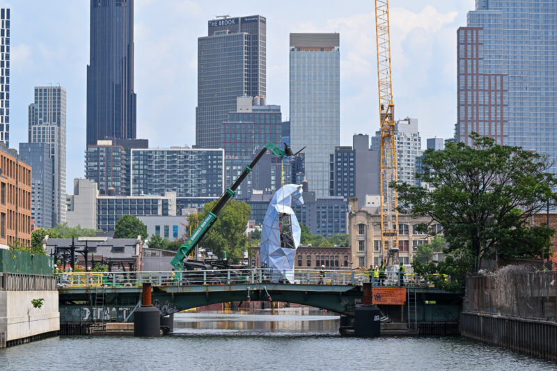 A metallic sculpture, Jen Lewin The Ursas, is being installed on a bridge over a canal in a city, with modern high-rise buildings and construction cranes in the background.