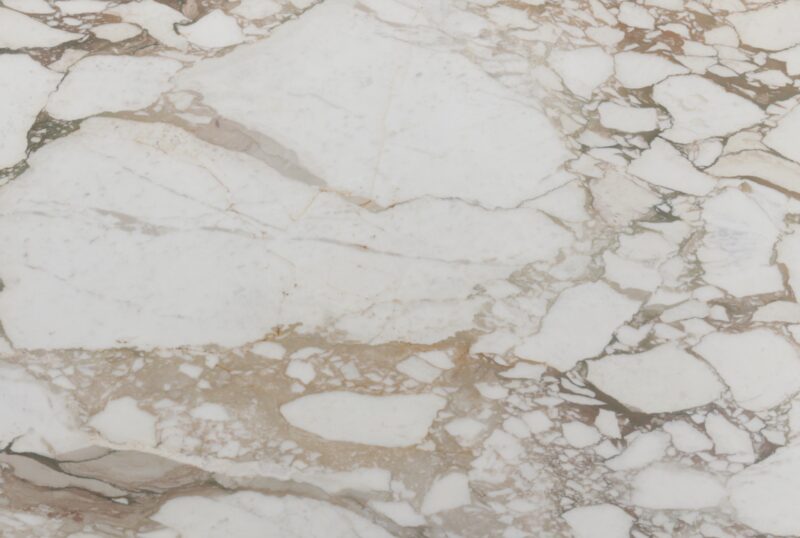 White marble surface with irregular brown and beige veining and patches, showing a natural stone texture and pattern, as curated by April Graves of Aria Stone Gallery.