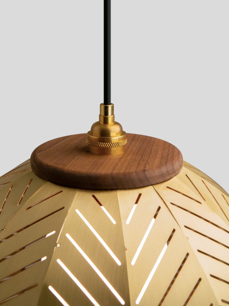 Close-up of a hanging pendant light, designed by Nick Sheridan for Cerno, with a gold metal shade featuring diagonal cutouts, topped with a wooden accent and brass fixture against a plain gray background.