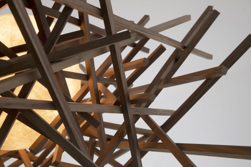 A close-up of a geometric wooden light fixture, designed by Nick Sheridan for Cerno, with interlocking wooden rods surrounding a glowing spherical light source.
